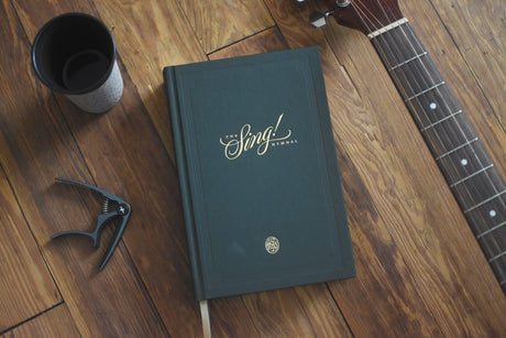 The Sing! Hymnal on a wooden table beside a guitar